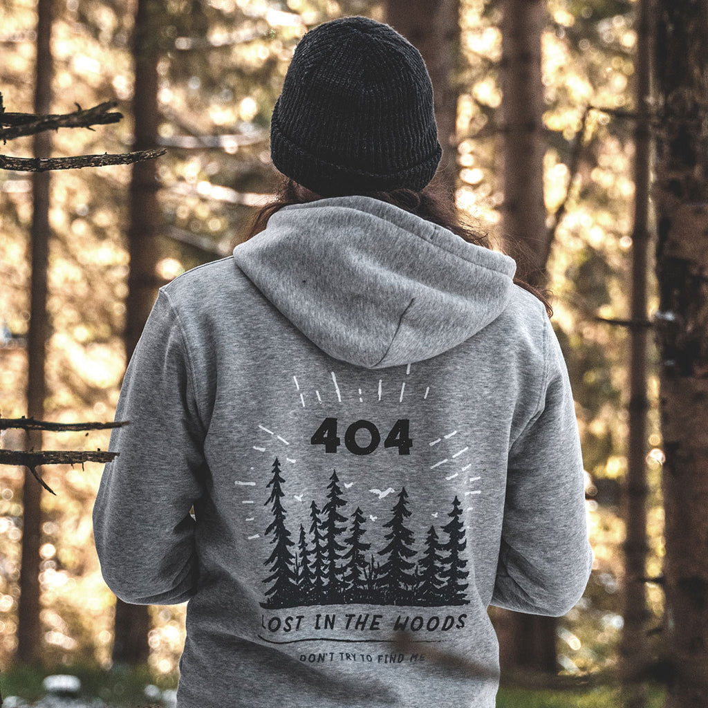 Model im Wald von hinten mit Harz Hoodie in Heather Grey mit blau-schwarzem Backprint: illustrierte Tannen und Text „404 – Lost in the woods – Don’t try to find me“. Nachhaltig produziert, Fair Wear & OEKO-TEX zertifiziert, pro Kauf ein Baum im Harz.