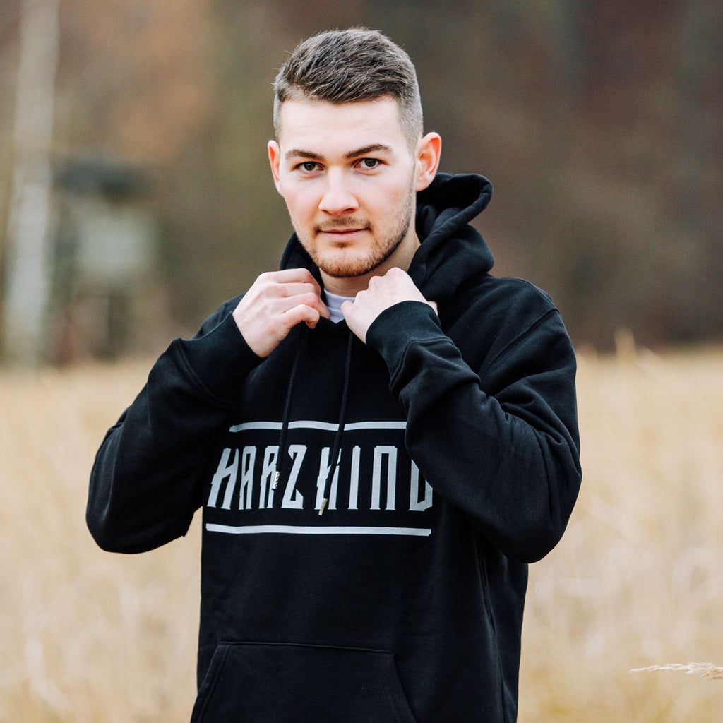 Männliches Model mit schwarzem Harz Hoodie mit weißem HARZKIND Logo – nachhaltig, Fair Wear & OEKO-TEX, pro Kauf 1 Baum im Harz